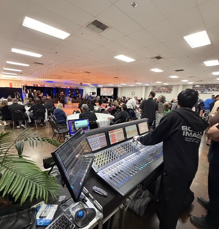 Avid S6L console at FOH during a black-tie gala — uplit cabaret tables and chandeliers visible past the meter bridge.