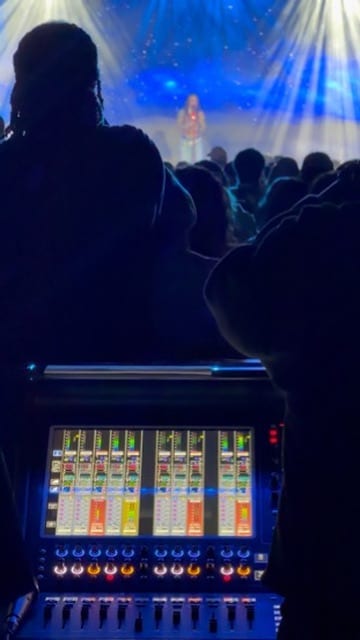 Over-the-shoulder from front-of-house — a digital console's channel meters glowing in the foreground, a singer on a blue-lit stage beyond a standing audience.