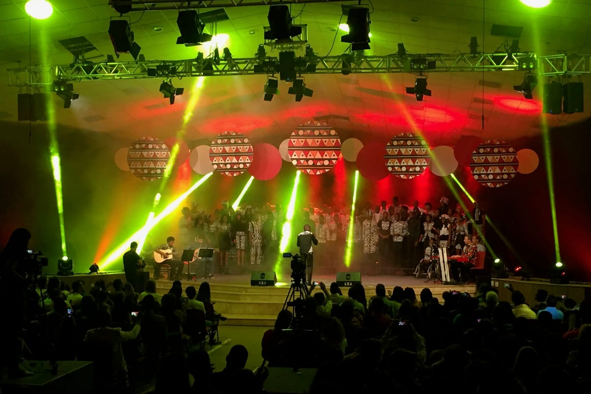 Wide concert frame — green and red laser beams cutting through the air, orange tribal-pattern backdrop discs glowing like planets, a full choir on risers between two broadcast camera silhouettes.
