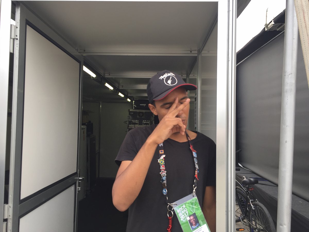 Victor backstage in a production trailer at Rock in Rio 2017, the festival's accreditation pass around his neck, gear down the corridor behind him.