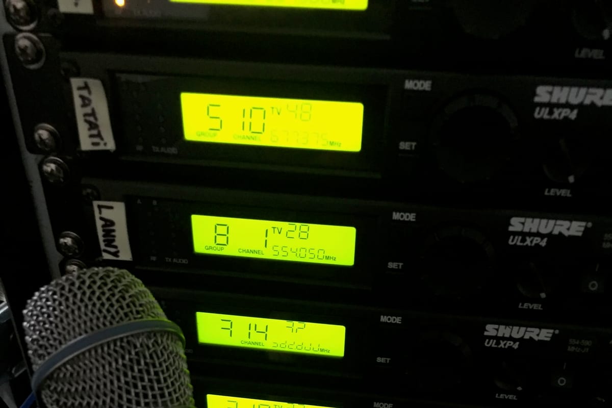 Close-up of a Shure ULXP4 wireless receiver rack — green LCDs showing group and channel frequencies, channels labeled on tape (TATAi, LANNY), a microphone capsule in the foreground.
