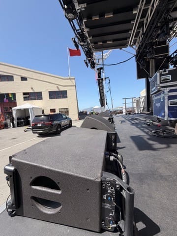 Festival stage deck mid-load-in — flight cases on the boards, line-array hangs in the background, crew moving cabling under house lights.