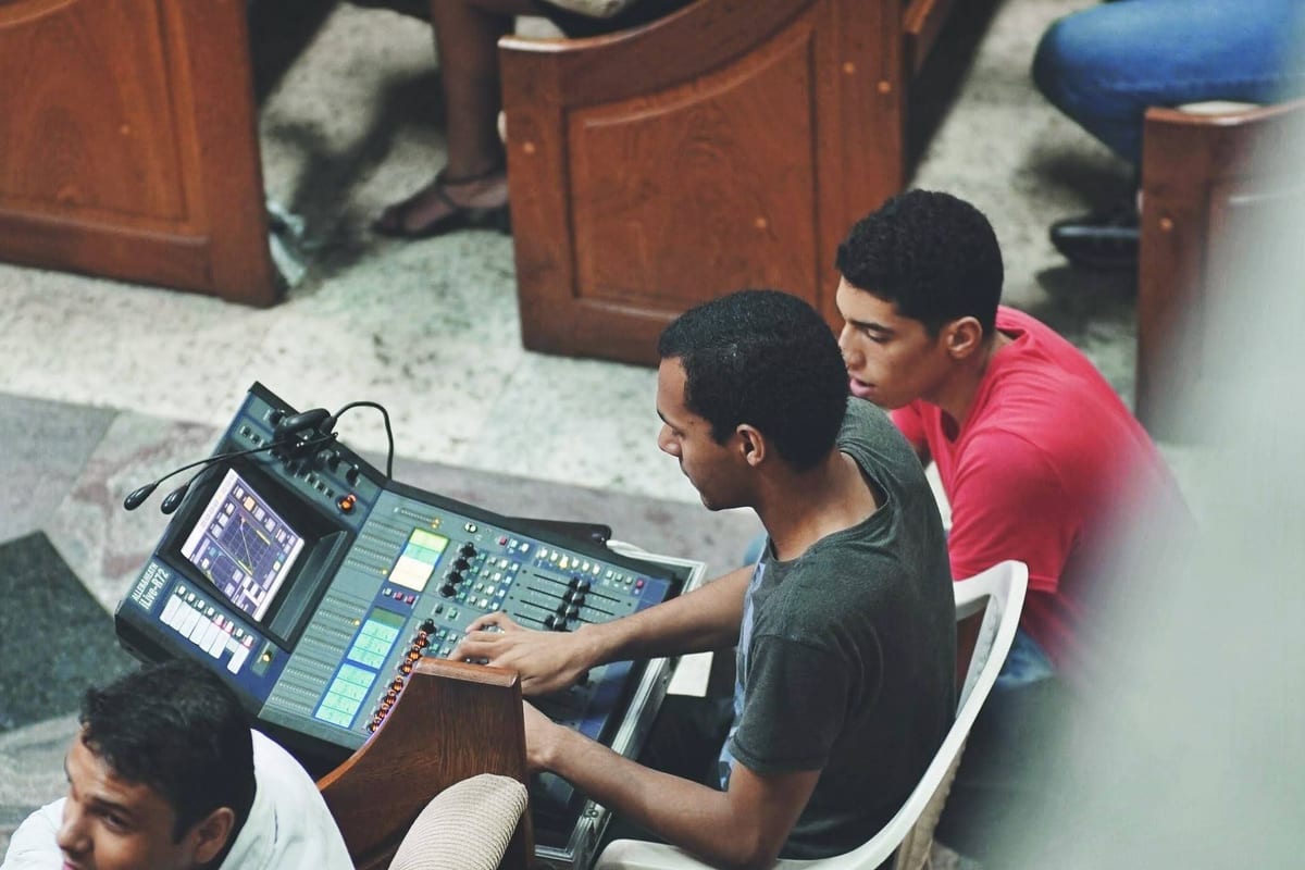 Bird's-eye view of Victor at a Roland V-Mixer, both hands on the console (touchscreen and knobs); a second engineer leans in.